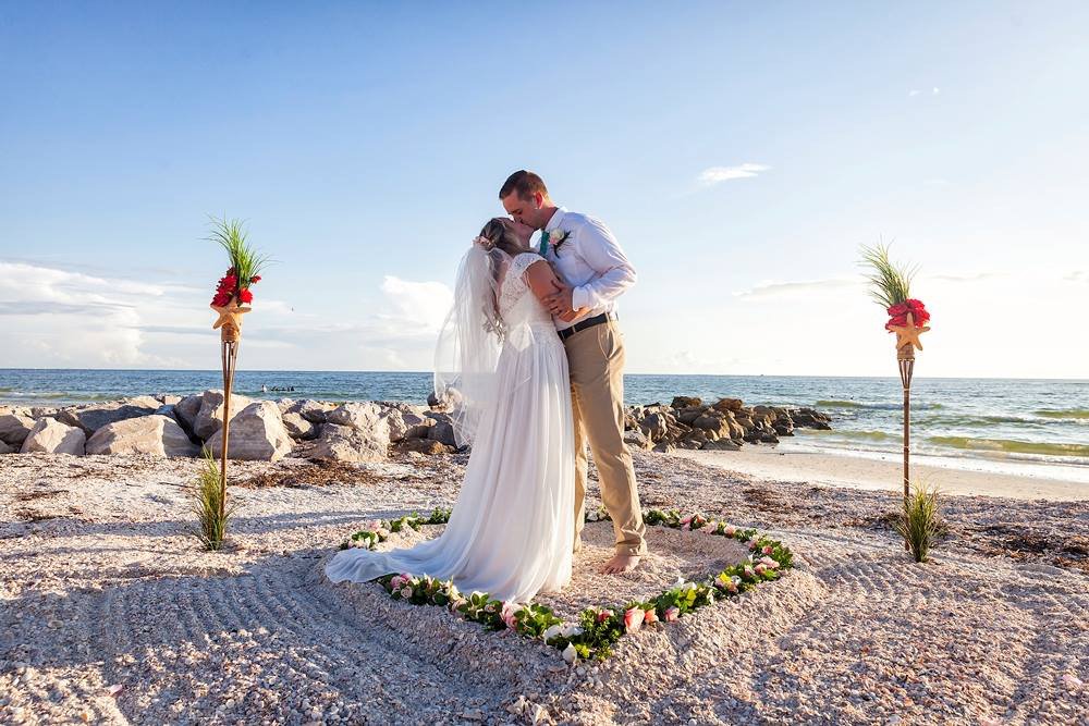 A bride and groom embracing on a shelly Gulf beach at Sunset Beach, a rocky jetty and the Gulf of Mexico behind them